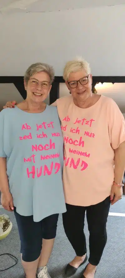 Zwei ältere Frauen tragen übergroße pastellfarbene **Hunde-Shirts** mit deutschem Text. Die Frau links trägt ein hellblaues Shirt und die Frau rechts ein hellrosa Shirt. Auf beiden Shirts steht ein leuchtend rosa Schriftzug „Hunde“. Sie stehen drinnen und lächeln in die Kamera.