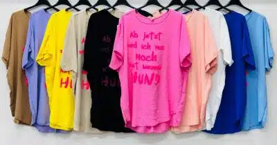 Eine Reihe bunter T-Shirts mit verschiedenen aufgedruckten Texten hängt an einem weißen Ständer vor einer weißen Wand. In der Mitte des rosa Hunde-Shirts steht in schwarzer Schrift „Ab jetzt red ich nur noch mit meinem HUND“.