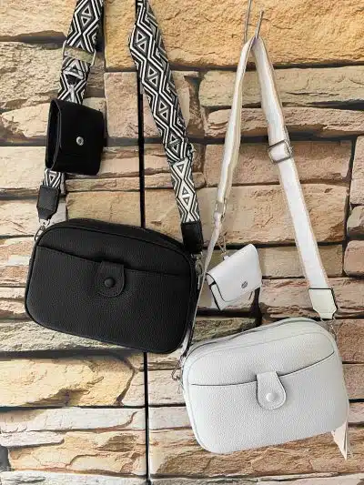 Mehrere kleine Moda Box Taschen mit unterschiedlichen Designs und Riemen hängen an einem Haken an einer Backsteinwand. Die Taschen variieren in Farbe und Muster, darunter auch schwarze und weiße Optionen.