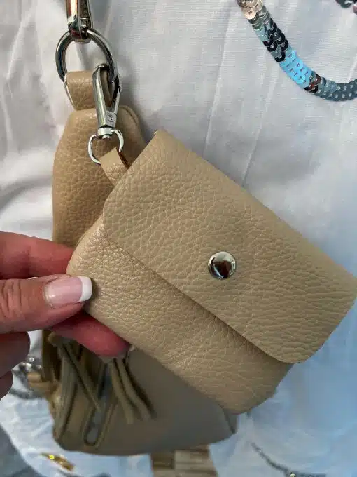 Nahaufnahme einer Hand, die eine kleine dreieckige Tasche aus beigefarbenem Leder hält, die an einem Schlüsselbund befestigt ist, mit einem Hintergrund aus hell gefärbtem Stoff und anderen Accessoires.