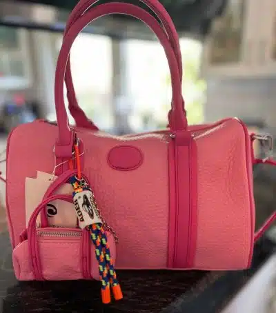 Eine pinkfarbene Style Bag mit Kieselstruktur und einem bunten Perlen-Schlüsselanhänger auf einer reflektierenden Oberfläche. Die Tasche hat einen oberen Griff, eine Außentasche und eine strukturierte Form.