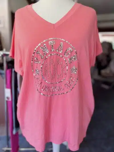 Ein rosa Vintage-Rock-Shirt mit V-Ausschnitt, präsentiert auf einer Schaufensterpuppe, mit einem Strass-Design in Form eines Kreises und den Worten „New York Original“ in der Mitte.