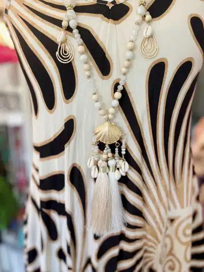 Eine Nahaufnahme einer mit weißen Perlen besetzten Liz-Boho-Kette mit einem großen Muschelanhänger und einer Quaste, präsentiert an einer Schaufensterpuppe, die ein Kleid mit Zebramuster trägt.