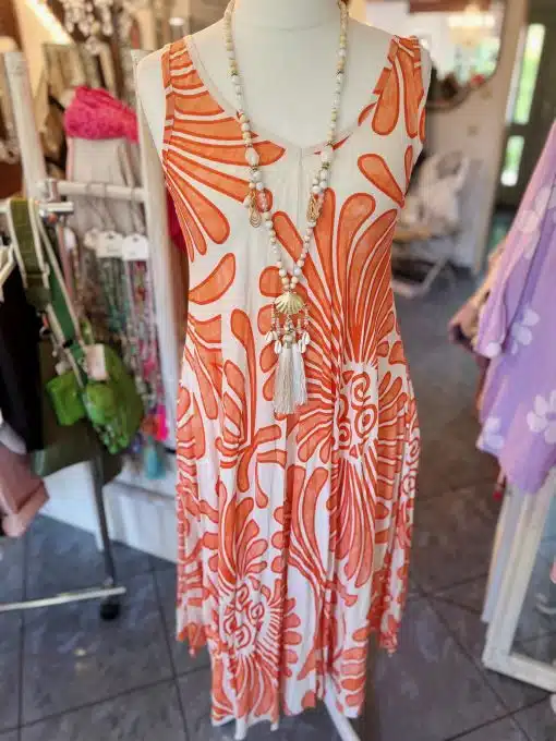 Ein ärmelloses, orange-weißes Sommerkleid von Iland mit Blumenmuster, ausgestellt auf einer Schaufensterpuppe in einer Boutique. Das Kleid ist mit einer langen Halskette mit Perlen und Quasten verziert.