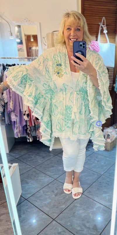 Eine lächelnde Frau macht ein Selfie in einem Spiegel und trägt dabei ein hellgrünes, fließendes Oberteil mit weißen Hosen und Sandalen. Im Hintergrund ist ein Bekleidungsgeschäft mit verschiedenen Kleidungsstücken zu sehen, in dem der Life Ibiza Boho Cardigan präsentiert wird.