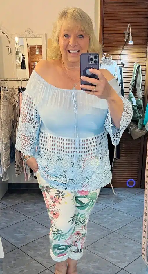 Eine Frau mit blondem Haar lächelt, während sie ein Selfie in einem Spiegel macht. Sie trägt eine weiß-blaue Ombre-Welcome-Tunika mit Spitzendetails und geblümte Hosen. Der Hintergrund zeigt einen Raum mit Kleidung und Spiegeln.