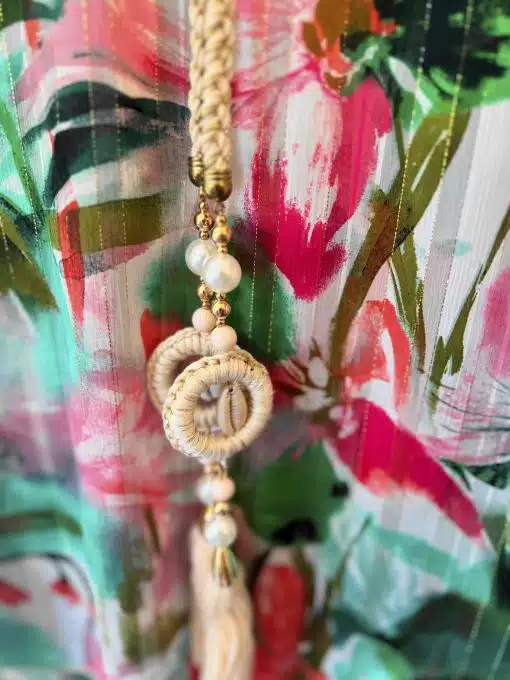 Eine Nahaufnahme einer dekorativen Quaste, die vor einem farbenfrohen Stoffhintergrund mit Blumenmuster hängt. Die Quaste weist geflochtene Details und Perlenverzierungen auf und verkörpert die Essenz des Paradies-Boho-Tunika-Stils.