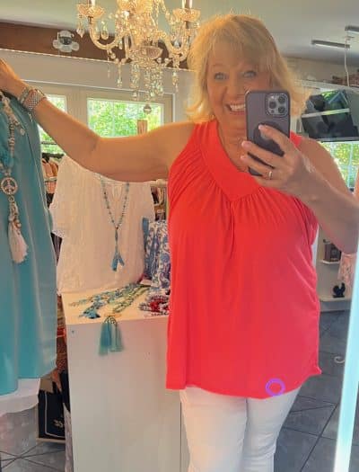 Eine Frau in einer leuchtenden Wassermelonen-Bluse aus Merinoseide und weißen Hosen macht ein Selfie vor einem Spiegel, umgeben von bunter Kleidung und Accessoires unter einem Kronleuchter.