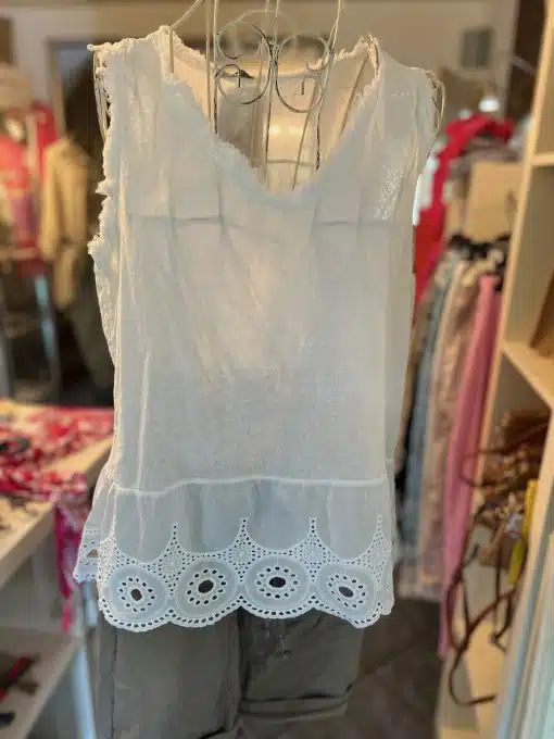 Ein weißes Romantik Boho Top wird auf einer Schaufensterpuppe ausgestellt. Das Boho Top hat einen V-Ausschnitt und einen gewellten Saum mit Ösendetails. Es hängt in einem Bekleidungsgeschäft, im Hintergrund sind verschiedene Kleidungsstücke zu sehen.