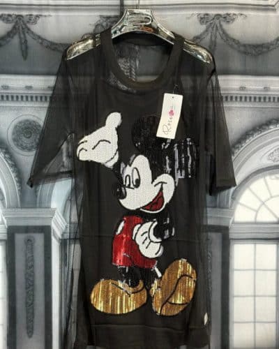 An einem Ständer hängt ein schwarzes Mickey-Netz-Shirt mit einem in Pailletten verzierten Mickey-Mouse-Bild auf der Vorderseite. Die Figur trägt rote Shorts und gelbe Schuhe. Am Ausschnitt des Shirts ist ein Etikett befestigt, das sich von einem Hintergrund mit einem kunstvollen Architekturmuster abhebt.
