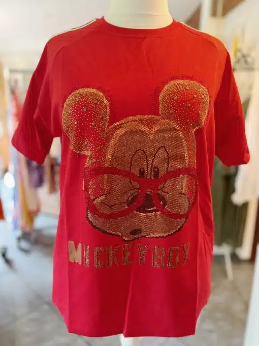 Ein rotes Mickey Boy-Shirt mit einem großen, glitzernden Design des Gesichts und der Ohren einer Cartoon-Maus auf der Vorderseite. Unter dem Gesicht steht das Wort „MICKEYBOY“ in glitzernden goldenen Buchstaben. Das Shirt wird auf einer Schaufensterpuppe in einem Raum mit verschwommenen Hintergrundgegenständen ausgestellt.