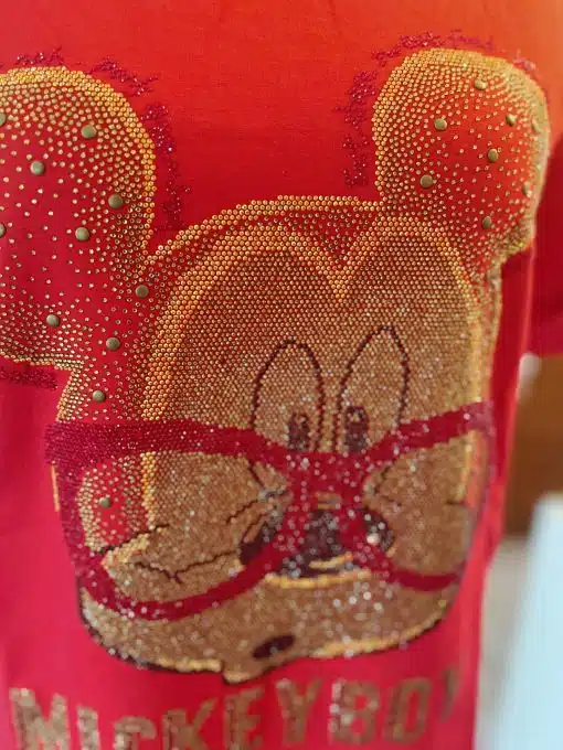 Nahaufnahme eines roten Mickey Boy-Shirts mit einem detaillierten Design des Gesichts einer Figur aus kleinen, bunten Perlen. Die Figur hat große Ohren, trägt eine Brille und unter dem Design steht der Name „Mickeyboy“.
