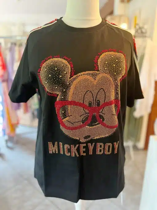 Ein schwarzes Hemd, das auf einer Schaufensterpuppe ausgestellt ist, zeigt eine große, glitzernde Grafik mit einem Cartoon-Mäusegesicht mit roter Brille. Unter der Grafik ist in Großbuchstaben das Wort „MICKEYBOY“ gedruckt. Dieses auffällige Mickey Boy-Shirt ist Teil einer Kleiderausstellung, im Hintergrund sind weitere Artikel zu sehen.