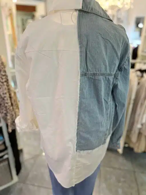 Eine Schaufensterpuppe trägt ein Hemd mit einem einzigartigen Design, das auf der linken Seite ein weißes langärmeliges Casual Jeans Hemd und auf der rechten Seite eine blaue Jeansjacke aufweist. Die Nähte sind asymmetrisch genäht, wodurch die beiden unterschiedlichen Stoffe miteinander verschmelzen. Umgeben sind verschiedene Freizeitkleidungsstücke und Jeans.