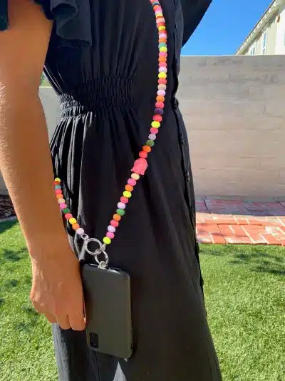 Eine Person in einem schwarzen Kleid hält eine Handy-Taschenkette, die an einem bunten Perlenband befestigt ist und wie eine modische Kette über der Schulter getragen wird. Im Hintergrund sind ein Ziegelweg und eine helle Wand unter dem klaren blauen Himmel zu sehen.
