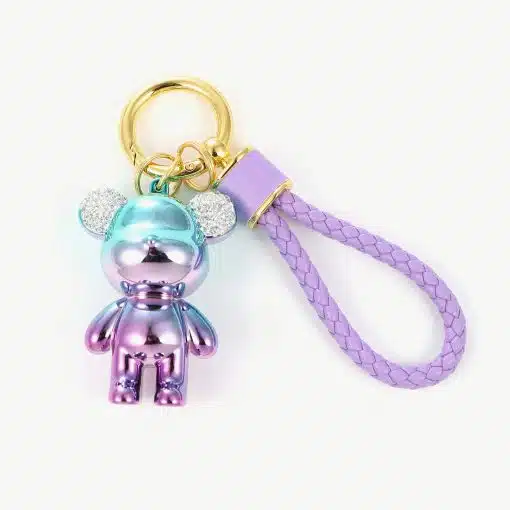Ein Schlüssel-Bär-Anhänger mit einem kleinen metallischen Bärenanhänger in einem Farbverlauf aus Blau, Lila und Rosa. Er hat glitzernde Ohren und ist an einem goldenen Schlüsselring mit einer lila geflochtenen Schlaufe aus Kunstleder befestigt.