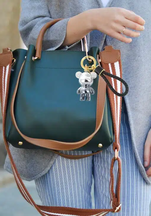 Eine Person hält eine grüne Handtasche mit braunen Riemen und goldenen Beschlägen. Die Tasche ist mit einem Schlüssel-Bär-Anhänger versehen. Die Person trägt einen grauen Pullover und blau-weiß gestreifte Hosen, die von der Taille abwärts sichtbar sind.