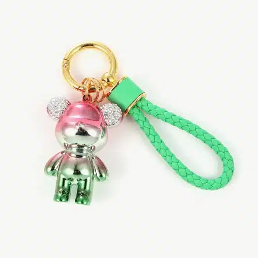 Ein Schlüssel-Bär-Anhänger mit einem metallischen Bären-Anhänger in Rosa- und Grünverlauf und glitzernden Ohren. Er ist an einem goldenen Ring und einer geflochtenen grünen Riemenschlaufe befestigt.