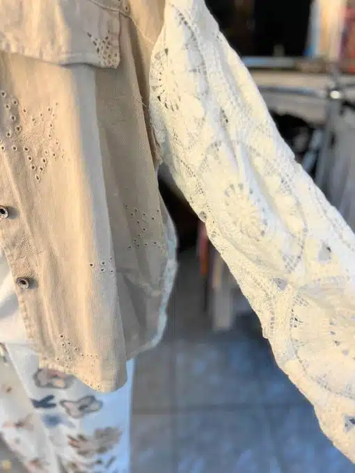 Eine Vintage Boheme Jeansjacke wird in einem Laden ausgestellt und verströmt Vintage-Charme. Der Ärmel ist verlängert und zeigt ein kontrastierendes weißes Spitzenteil. Die Regale und Ständer mit anderen Kleidungsstücken sind im unscharfen Hintergrund sichtbar.