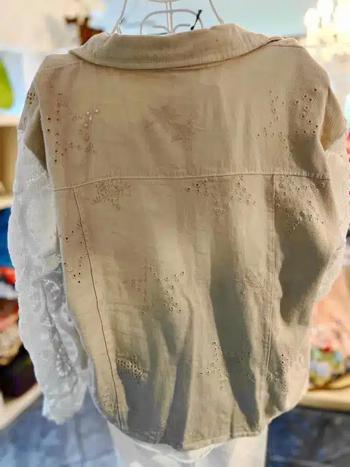 Eine Vintage-Boheme-Jeansjacke mit gestickten Blumenmustern auf der Rückseite wird auf einer Schaufensterpuppe präsentiert. Die Jacke, die einen Boheme-Charme ausstrahlt, weist Ösendetails auf und ist mit einem Ärmel aus weißer Spitze kombiniert. Im Hintergrund sind ein Regal mit verschiedenen Gegenständen und ein Kronleuchter zu sehen.