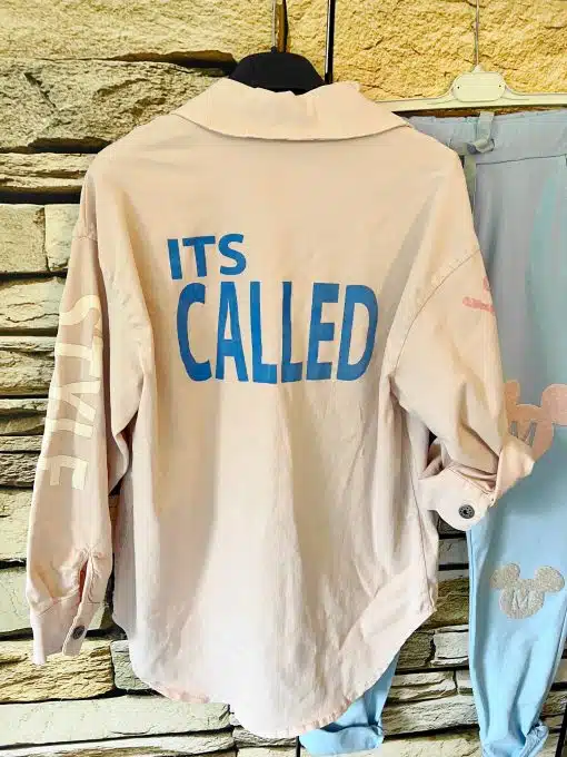 Ein hellrosa Hemd mit Knopfleiste hängt an einem Ständer vor einer Steinmauer. Auf dem Rücken des Hemdes steht in kräftigem Blau „IT’S CALLED“. Daneben ergänzt eine It’s Call-Jeansjacke mit teilweise sichtbaren hellblauen Grafiken das Ensemble.