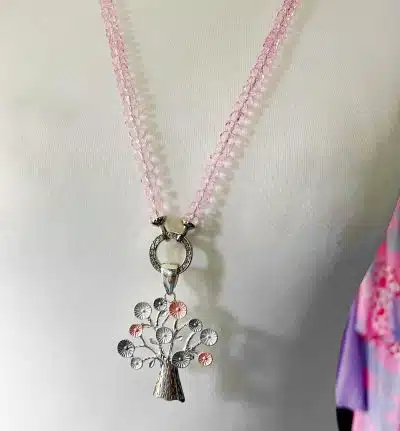 Eine rosa Perlenkette mit einem silbernen Wunschbaum-Wechselanhänger in Form eines Baums wird auf einem weißen Puppentorso präsentiert. Der aufwendig gestaltete Anhänger zeigt zarte Zweige und Blätter. Der Hintergrund ist mit einem Hauch von rosa und blauem Stoff versehen.