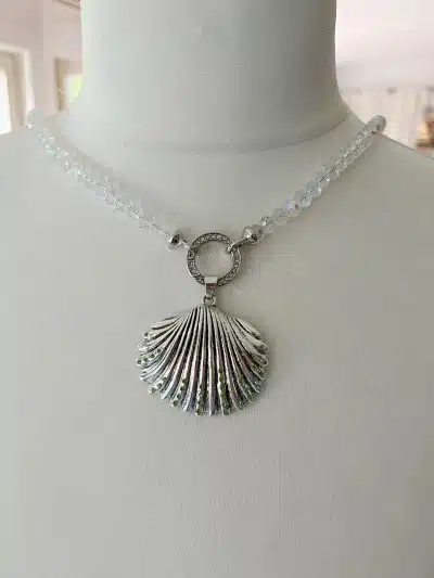 Eine weiße Schaufensterpuppe präsentiert eine Halskette aus klaren, runden Perlen. Die Halskette verfügt über einen großen, silberfarbenen Muschel-Wechselanhänger mit einem komplizierten Design, der durch einen runden, silberfarbenen, mit kleinen Strasssteinen verzierten Verbinder mit den Perlen verbunden ist.