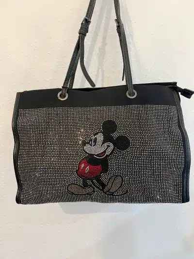 Eine schwarze Mickeys City Tasche mit zwei Henkeln ist mit einem Bild von Mickey Mouse aus kleinen, funkelnden Strasssteinen verziert. Mickeys ikonische rote Shorts heben sich wunderschön von der schlichten weißen Wand ab und machen diese Tasche perfekt für einen schicken Stadtbummel.