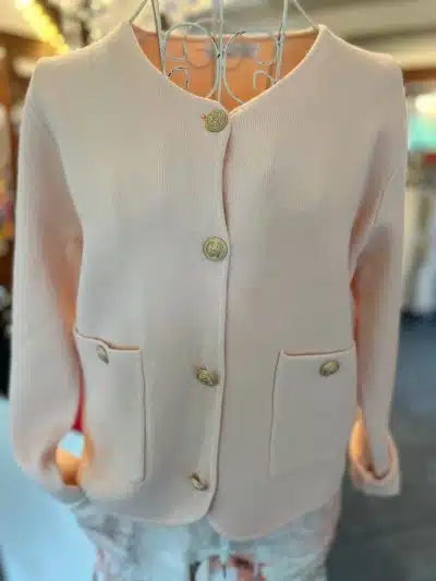Auf einem Kleiderbügel hängt eine hellrosa, langärmelige, zugeknöpfte Short Jacke. Die Strickjacke im Stil einer kurzen Jacke hat einen runden Ausschnitt, zwei Vordertaschen und goldfarbene Knöpfe.