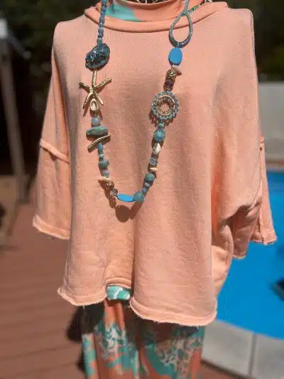 Eine Vintage-Schaufensterpuppe trägt ein Uni-Vintage-Sweatshirt mit langen Ärmeln über einer Hose mit aqua- und orangefarbenem Muster. Das Outfit wird durch eine lange, klobige Halskette ergänzt, die mit verschiedenen blauen Perlen und einem Seesternanhänger geschmückt ist.