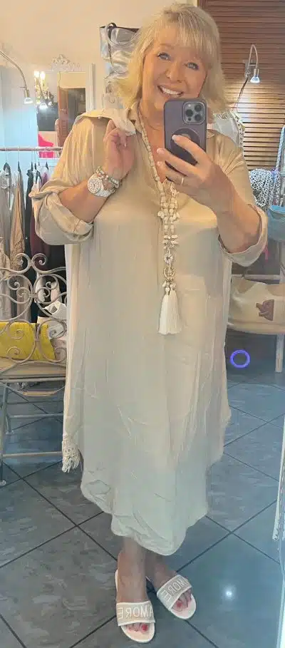 Eine Frau mit hellem Haar macht ein Selfie vor einem Spiegel. Sie trägt ein Edel Dress Kleid, eine lange Kette mit Quasten und weiße Pantoletten mit der Aufschrift „AMORE“. Im Hintergrund ist ein Raum zu sehen, in dem verschiedene Stoffe und Kleidungsstücke ausgestellt sind.