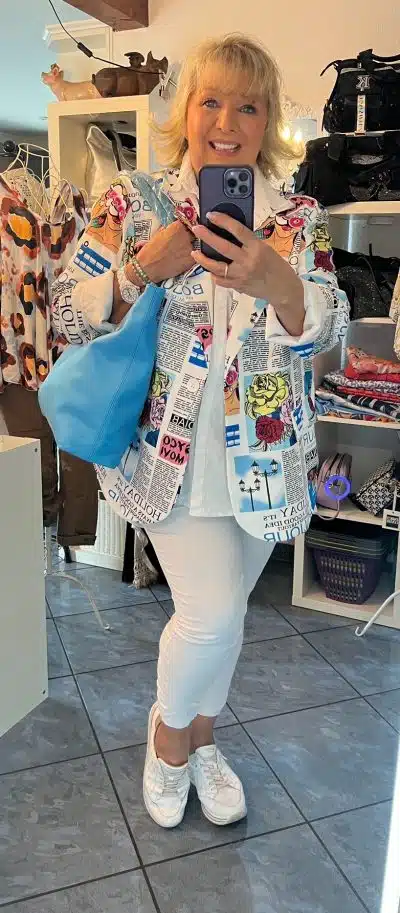 Eine Person steht vor einem Spiegel und macht mit einem Smartphone ein Foto. Sie trägt ein weißes Outfit mit einem Blazer XL aus der Zeitungsausgabe, weiße Turnschuhe und eine blaue Tasche. Im Hintergrund sind Regale mit Kleidung und Accessoires zu sehen.
