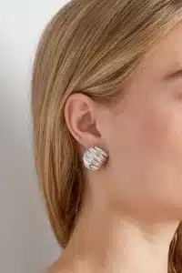 Eine Nahaufnahme des Ohrs einer Frau, geschmückt mit einem großen, runden, strukturierten Basic-Ohrstecker in Silber. Das glatte blonde Haar der Frau bedeckt teilweise ihr Ohr und verleiht dem Look eine dezente Eleganz. Der Hintergrund ist schlicht und unscharf