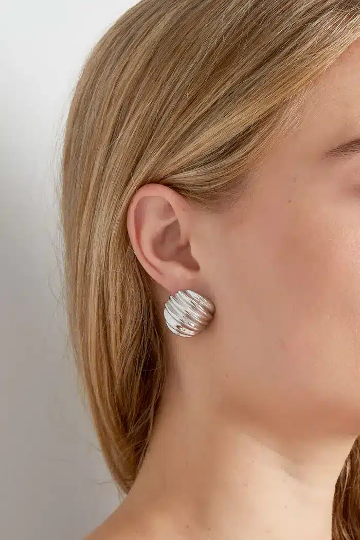 Eine Nahaufnahme des Ohrs einer Frau, geschmückt mit einem großen, runden, strukturierten Basic-Ohrstecker in Silber. Das glatte blonde Haar der Frau bedeckt teilweise ihr Ohr und verleiht dem Look eine dezente Eleganz. Der Hintergrund ist schlicht und unscharf.