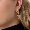 Nahaufnahme des Ohrs einer Person, die mit exquisiten Love-Ohrsteckern geschmückt ist. Das geometrische Ohrhänger-Design weist eine dreieckige Form auf, die mit einer rechteckigen Form mit aufwendigen Aussparungsdetails verbunden ist. Die Person mit hellbraunem Haar und einem schwarzen Rollkragenpullover strahlt eine Aura der Eleganz und eine Liebe für schicke Accessoires aus.