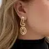 Nahaufnahme einer Person, die den Love-Ohrstecker trägt, einen goldenen Ohrring in Form des Buchstabens „L“, der über einem kreisförmigen Design hängt. Der Kopf der Person ist leicht zur Seite gedreht und zeigt blondes Haar und einen Teil eines schwarzen Rollkragenpullovers. Eine goldene Halskette verleiht einen zusätzlichen Hauch von Liebe und ergänzt den eleganten Love-Ohrstecker perfekt.