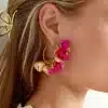 Nahaufnahme des Ohrs einer Person, das mit großen, dekorativen Herbstblumen-Ohrsteckern in Gold- und leuchtenden Rosatönen geschmückt ist. Die Person hat langes, blondes Haar, das mit einer goldenen Haarspange befestigt ist, und der Saum eines beigen Kleidungsstücks ist ebenfalls sichtbar.