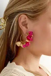 Nahaufnahme des Ohrs einer Person, das mit großen, dekorativen Herbstblumen-Ohrsteckern in Gold- und leuchtenden Rosatönen geschmückt ist. Die Person hat langes, blondes Haar, das mit einer goldenen Haarspange befestigt ist, und der Saum eines beigen Kleidungsstücks ist ebenfalls sichtbar