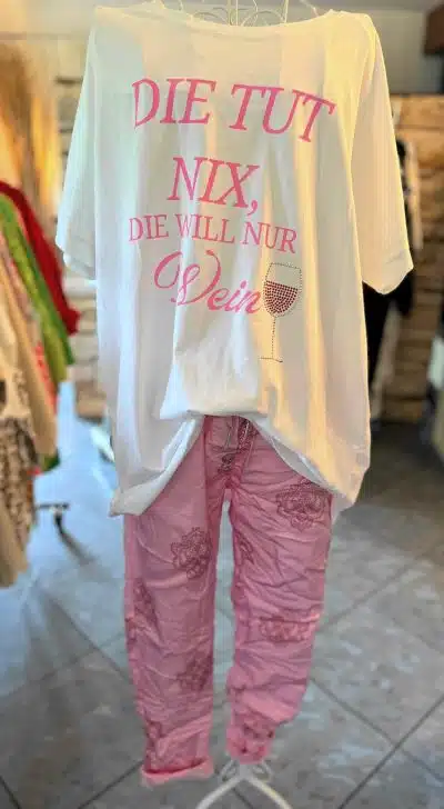 Ein weißes „Die tut nix“-Shirt mit pinkfarbenem deutschen Text, der übersetzt „Sie tut nichts, sie will nur Wein“ bedeutet, wird auf einem Ständer ausgestellt. Dazu passen pinkfarbene Hosen mit Blumenmuster. Die Kleidung wird in einem Geschäft präsentiert, im Hintergrund sind andere Kleidungsstücke zu sehen.