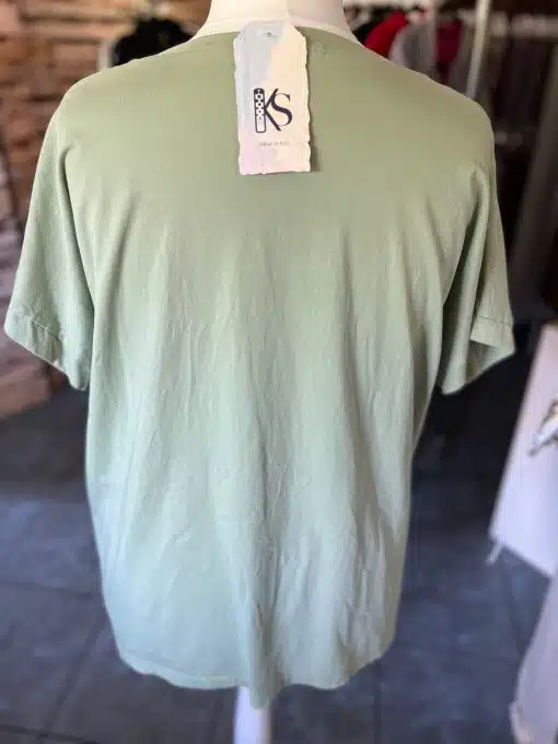 Ein hellgrünes kurzärmeliges Vintage-Rock-Shirt wird auf einer Schaufensterpuppe präsentiert. Am Kragen des Shirts hängt ein Schild mit der Marke „Kimes Ranch“. Im Hintergrund ist ein verschwommenes Interieur mit verschiedenen Kleidungsstücken und Holztexturen zu sehen.