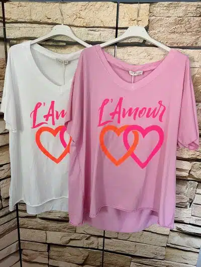 Zwei T-Shirts hängen an weißen Plastikbügeln vor einer Steinmauer im Hintergrund. Beide Shirts zeigen den Text „L'Amour“ und überlappende Herzgrafiken. Ein Làmour-Shirt ist weiß und das andere ist rosa.