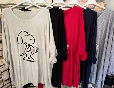Fünf langärmelige Snoopy Light Pullis hängen an weißen Kleiderbügeln. Die Shirts gibt es in verschiedenen Farben: Hellgrau mit Snoopy, der ein Herz hält, Schwarz, Rot, Marineblau und Dunkelgrau. Sie sind vor einer Wand mit Steinmuster ausgestellt und fangen das Licht wunderschön ein.