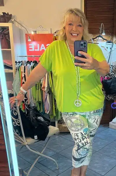 Eine Person mit blonden Haaren lächelt und macht ein Spiegel-Selfie in einem Bekleidungsgeschäft. Sie trägt ein XL-hellgrünes Basic-V-Shirt, gemusterte Hosen und eine lange Halskette mit einem Friedenszeichen. Im Hintergrund hängen verschiedene Kleidungsstücke und ein rotes Verkaufsschild ist sichtbar.