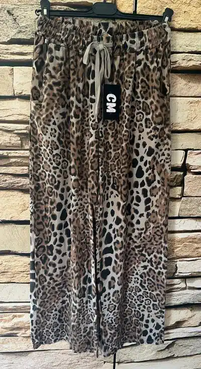 Eine Wildcat Chic Hose hängt vor einer Backsteinmauer. Die Hose mit Leopardenmuster hat einen elastischen Bund mit Kordelzug und ein Etikett mit der Aufschrift „CM“.