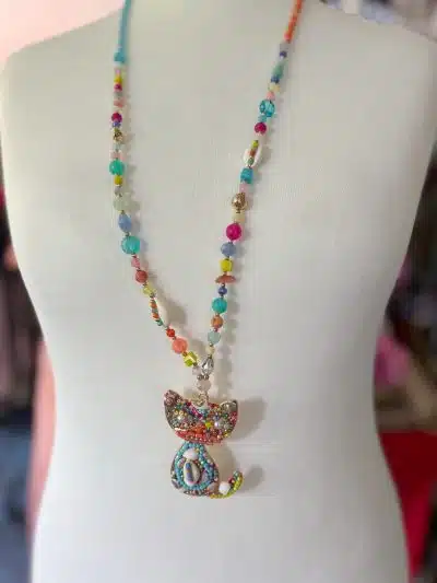 Eine Schaufensterpuppe präsentiert die „Cat Boho Kette“, eine Halskette im Bohème-Stil, die mit einer Reihe bunter Perlen verziert ist und einen filigranen Anhänger in Katzenform aufweist, der mit verschiedenen kleinen, leuchtenden Steinen und Juwelen verziert ist.