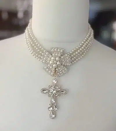 Das Royal Chic Collier ist eine mehrreihige Perlenkette mit einem großen dekorativen Verschluss, der mit einer Perlenansammlung und aufwendigen Strasssteinen verziert ist. Das raffinierte Design wird durch einen mit Edelsteinen verzierten Kreuzanhänger in der Mitte noch weiter aufgewertet, der dem Gesamtbild Eleganz verleiht.