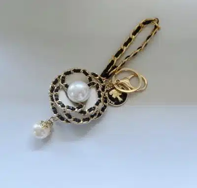 Der Bloom Key Charm ist ein eleganter Schlüsselanhänger, der mit einer großen, perlenartigen Perle in der Mitte eines kreisförmigen Webmusters verziert ist, neben der eine kleinere perlenartige Perle baumelt. Dieser dekorative Anhänger verfügt außerdem über mehrere goldene Ringe und ein schwarz-goldenes Kettenband.