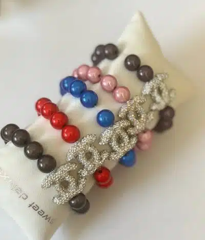 Auf einem weißen Kissen sind vier Magic Beads CC-Armbänder in Schwarz, Rot, Blau und Pink zu sehen. Darüber hinaus ist eine silberfarbene, mit Strasssteinen besetzte Metallkettenversion des CC-Armbands elegant mit den Perlenarmbändern verflochten.