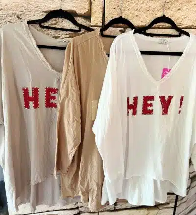 Drei Hey Vintage-Hemden hängen lose auf schwarzen Kleiderbügeln an einer strukturierten Wand. Die Hemden sind beige, braun und weiß. Auf den beigen und weißen Hemden steht in großen roten Buchstaben „HEY!“, während auf dem braunen Hemd in roten Buchstaben „HE“ steht.