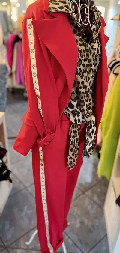 Eine Schaufensterpuppe präsentiert ein stylisches Outfit, bestehend aus einem roten Hosenanzug und einer Bluse mit Leopardenmuster. Der Anzug hat einen weißen Seitenstreifen mit einem Logomuster. Das Ensemble steht in einem hell erleuchteten Bekleidungsgeschäft.